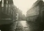En regnvåt gata i Ystad (?) med korsvirkeshus m.m. På en skylt till höger kan man läsa: 