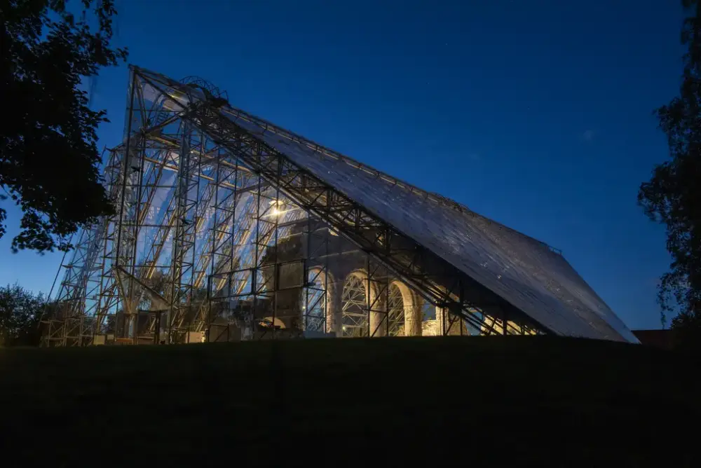 Kveldsbilde av svakt opplyst domkirkeruin (søylerekke med romanske buer) inne i Hamardomen, som er et vernebygg i glass og stål.