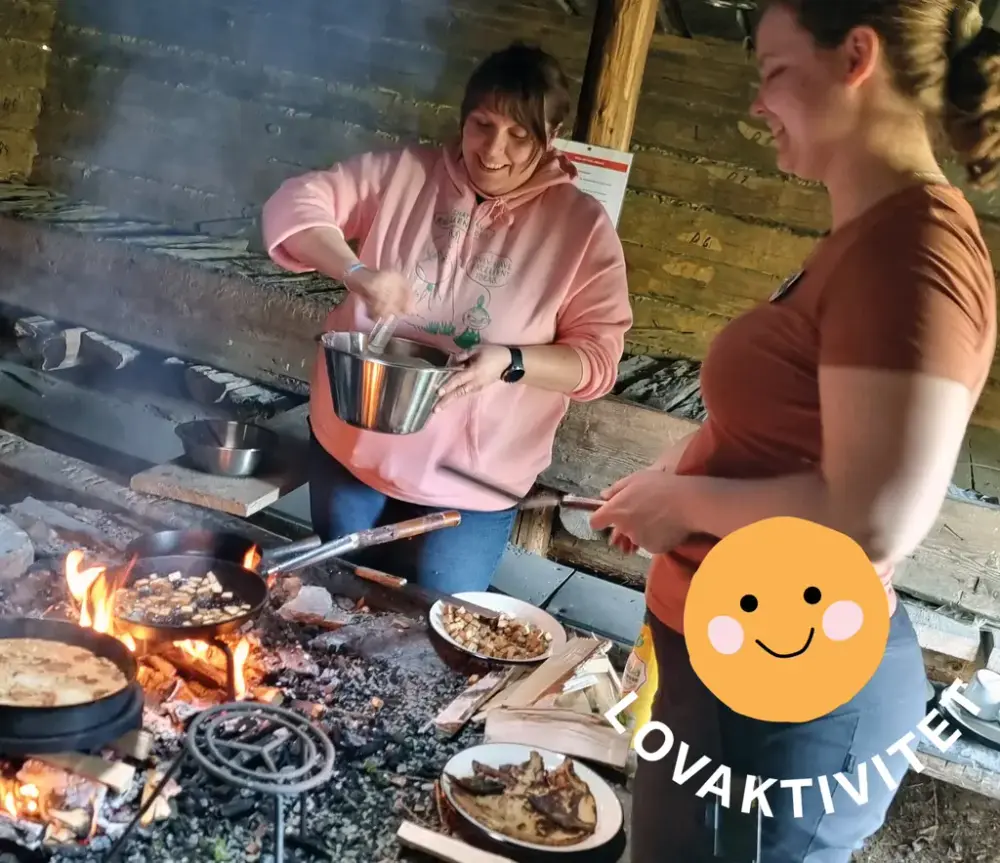 Två kvinnor står vid öppen eld och lagar mat samt tallrikar med mat på.