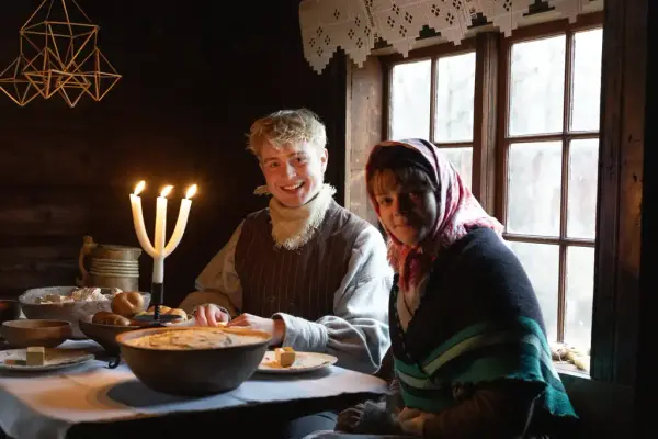 Smilende tunverter i gamle kostymer ved et bord med grøt, brød og tre lys. Strå-julepynt henger fra taket.