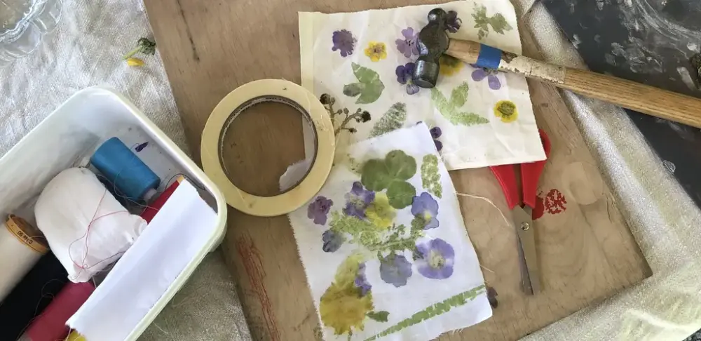 Bild tagen uppifrån. Tygbitar med blommönster, en hammare, en sax, tejp och en plastbytta med sytråd.
