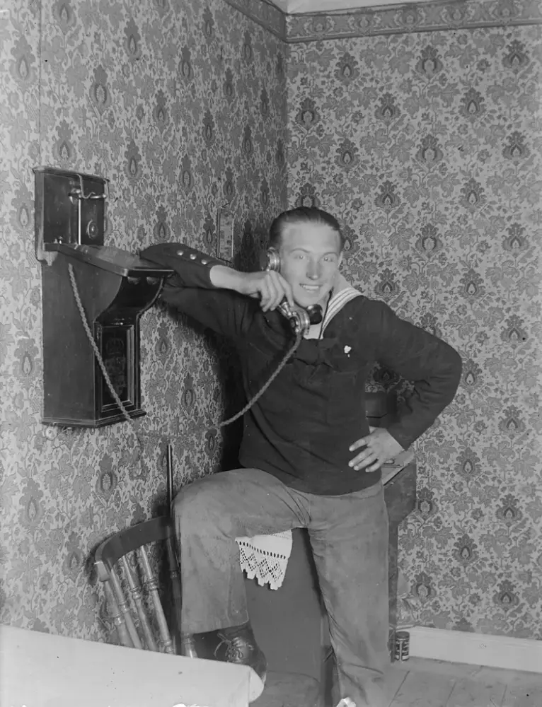 En man pratar i en gammeldags telefon och har högerbenet uppvräkt på ryggstödet av en stol.