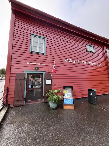 Vi ser den røde fasaden til Norges Fiskerimuseum, med inngangsdør i glass og brune karmer. Det er blomst utenfor, samt en reklamebukk og et bosspann