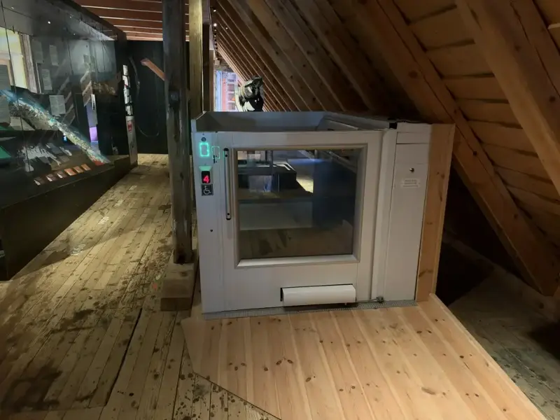 Bildet viser en heis, i utstillingen ressurser til låns. Det er en lav dør, som fører til en åpen heis og den går kun nedover til etasjen under. 