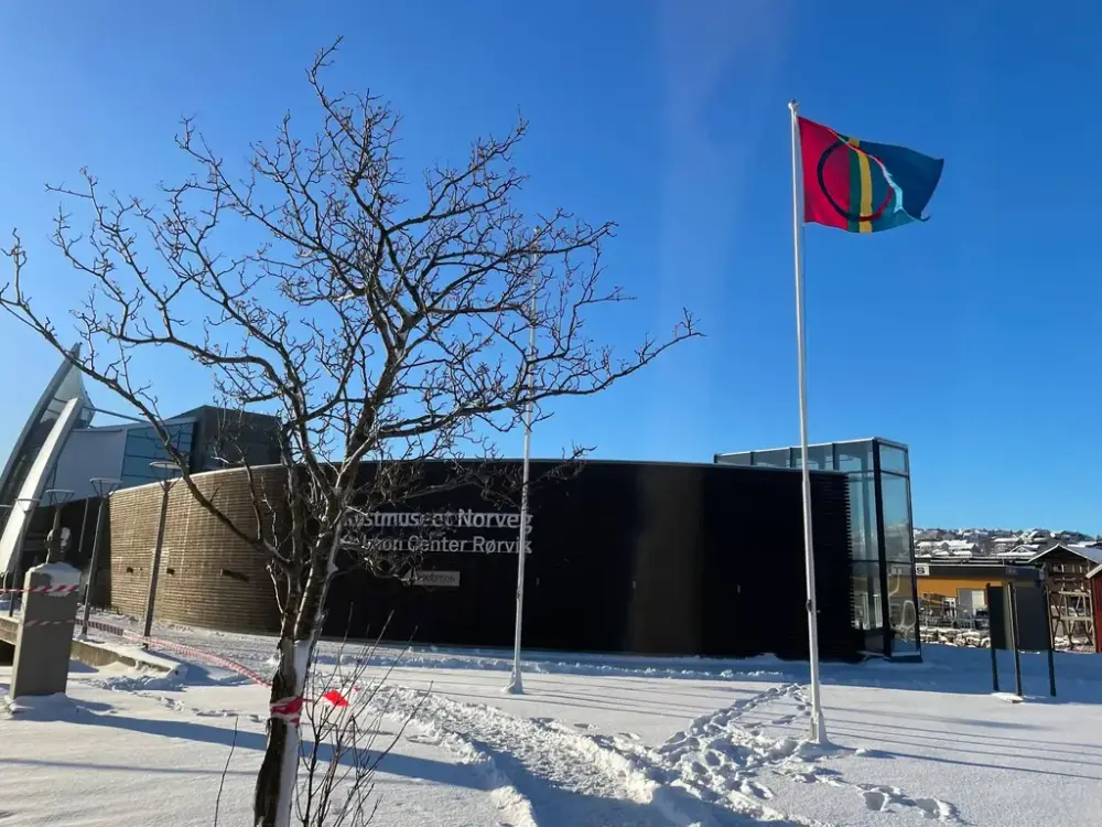 Samisk flagg er heist utenfor Kystmuseet Norveg med blå himmel og snø på bakken.